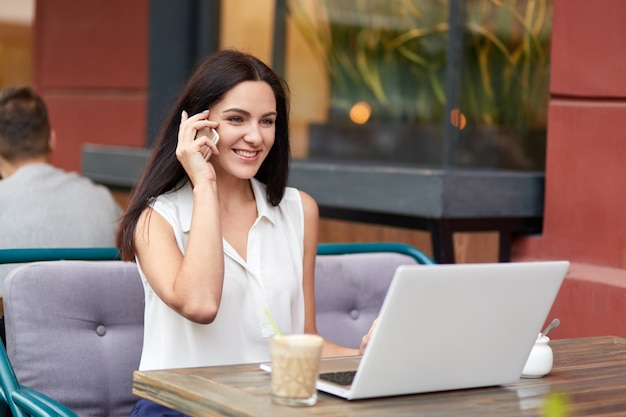 Positive businesswoman in formal clothes, has telephone conversaton, speaks with colleague, uses laptop computer for making business report