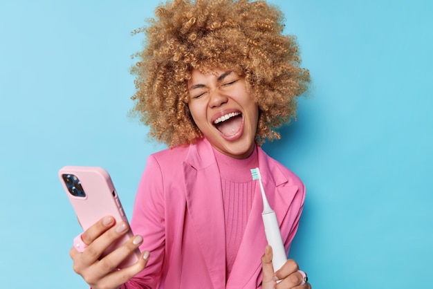 Positive carefree woman with curly bushy hair holds electric brush as if microphone makes selfie photo via smartphone dressed in formal pink jacket isolated over bue background Hygiene procedures