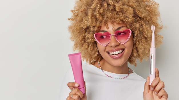 긍정적인 평온한 젊은 여성은 치약 튜브를 들고 전동 칫솔은 정기적으로 분홍색 하트 모양의 선글라스와 티셔츠를 흰색 배경의 빈 복사 공간에 격리되어 있습니다