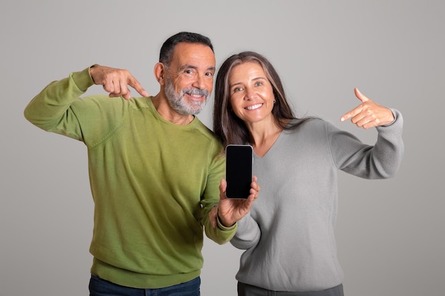 Positive caucasian aged lady and husband show fingers on smartphone advice app social network