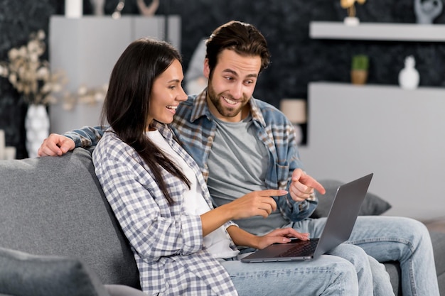 Positive caucasian couple using laptop while resting on couch\
at living room at home spending time together browsing social media\
internet watches videos messaging with friends smiling