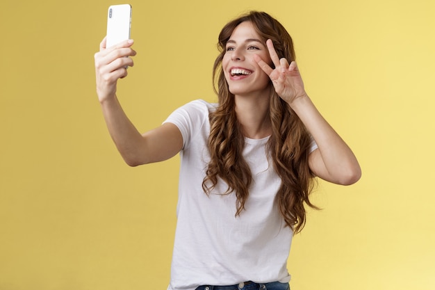 Positive charisamtic happy young girl curlyhaired raise smartphone taking selfie show peace victory ...