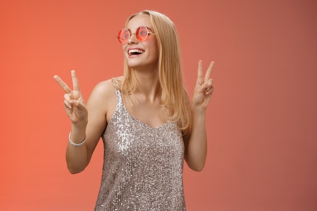 Positive charming european carefree stylish girlfriend having fun dance-floor show victory peace signs look up happily smile in sunglasses glamour glittering dress enjoy celebration, red background