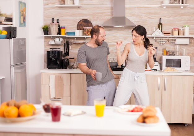 - Positive cheerful crazy couple dancing while having breakfast in kitchen wearing pajamas. Carefree wife and husband laughing having fun funny enjoying life authentic married people positive happy re