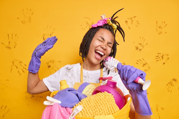 Positive cheerful housewife sings song along holds brush as if microphone wears dirty clothes stands near laundry basket wears rubber gloves foolishes around yellow wall