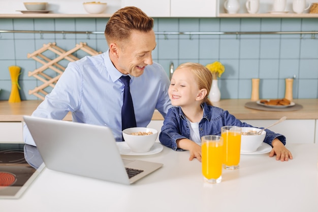 Positive cheerful nice girl looking a the laptop screen and smiling while having breakfast together with her father