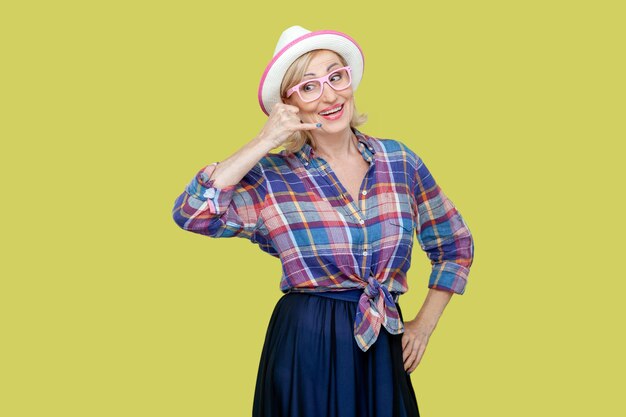 Positive cheerful senior woman in shirt hat and eyeglasses makes phone gesture says call me back