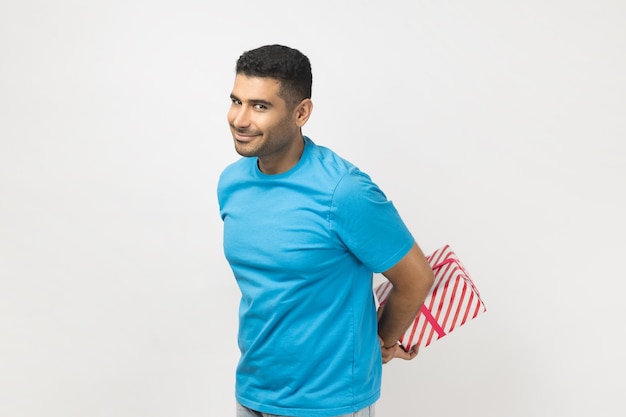 Positive cheerful unshaven man with present box behind back gift for holiday congratulating