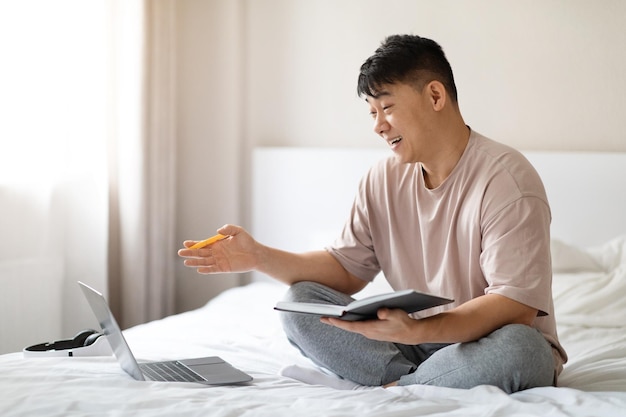 Positive chinese man using laptop in bed at home