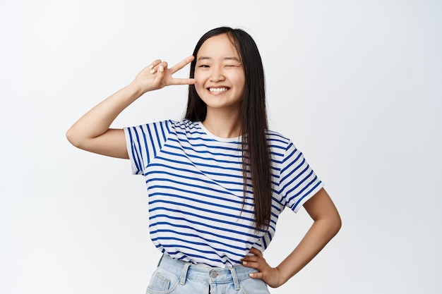 Positive and confident asian girl winking, smiling and showing happy peace sign near eye, kawaii pose, standing on white.