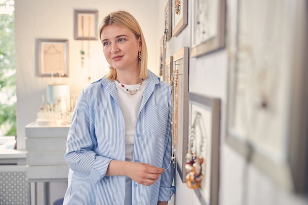 Positive consultant peacefully looking away while standing near store wall with necklaces