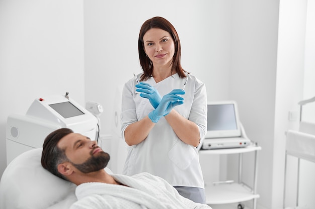 Positive cosmetologist holds syringes with filler for skin lifting standing by mature man in salon