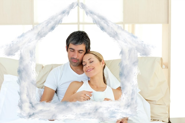 Positive couple drinking coffee lying in the bed against house outline in clouds