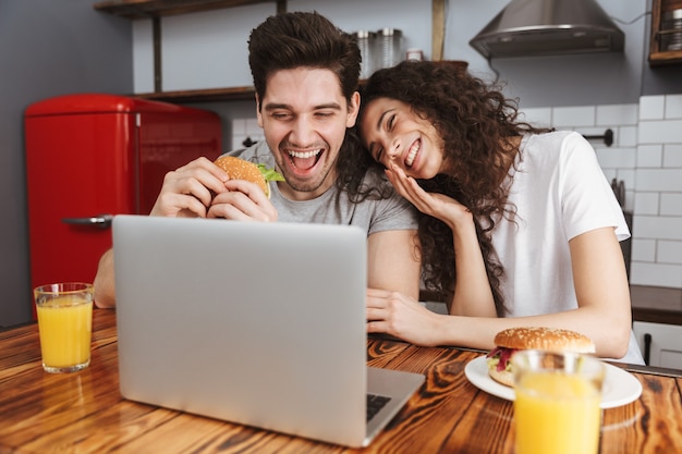 positive couple man and woman looking at laptop on table while eating hamburger in kitchen at home