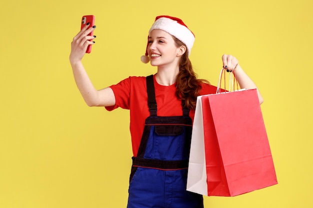 Positive courier female with smile taking selfie or having video call while holding shopping bags, wearing blue overalls and santa claus hat. Indoor studio shot isolated on yellow background.