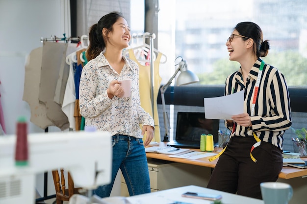 Positive crew of female fashion designers laughing discussing clothing sketches in studio. cheerful girls coworkers looking on illustration talking on new ideas on textile while drinking coffee