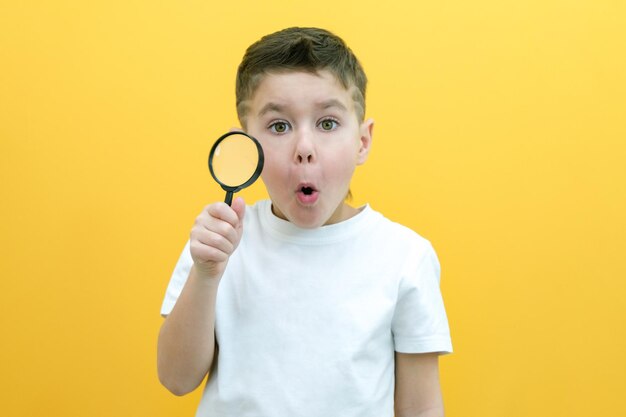 Positive curious schoolboy in casual clothes looks at the camera through a magnifying glass on a yellow background surprise