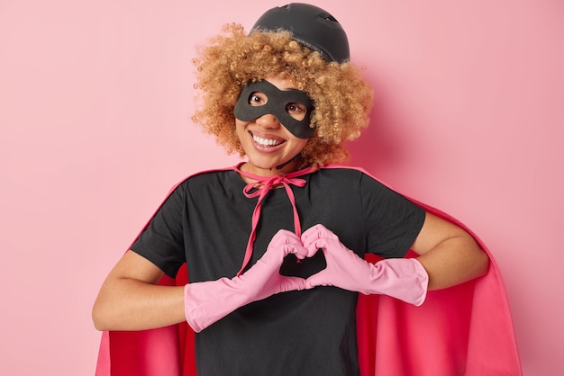 Positive curly haired female superhero being in love with\
someone makes heart sign with hands smiles positively wears\
superhero costume rubber gloves isolated over pink background be my\
valentine
