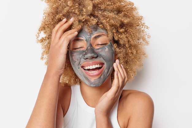 Positive curly haired woman keeps eyes closed applies grey clay mask touches face and giggles happily dressed casually has bare shoulders isolated over white background. Beauty treatments concept