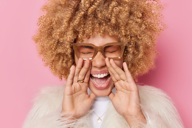 Positive curly haired woman keeps palms near face exclaims loudly closes eyes shouts or calls someone wears spectacles winter coat isolated over pink background. Human emotions and feelings.