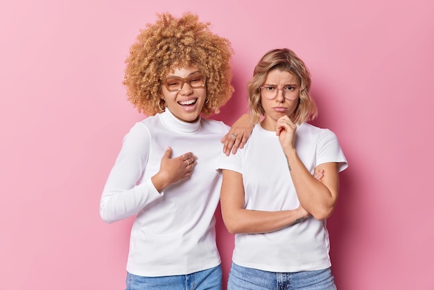 Positive curly haired woman laughs joyfully leans at shoulder of her sad friend dressed casually isolated on pink background People relationship and emotions concept Unhappy female model has problem