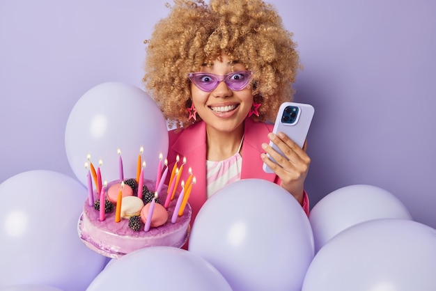 Positive curly haired woman smiles happily holds smartphone\
invites friends to birthday party holds tasty sweet cake with\
burning candles surrounded by inflated balloons isolated on purple\
background