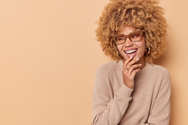 Positive curly woman looks joyfully away keeps hand on chin smiles broadly shows white teeth feels very glad wears transparent eyeglasses and jumper isolated over beige background blank space