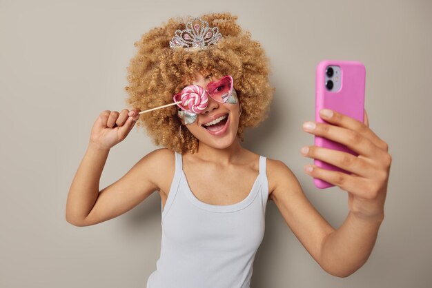 Positive curly woman wears small crown on head and casual t\
shirt covers eye with lollipop smiles joyfully clicks selfie via\
smartphone applies beauty patches under eyes poses against grey\
background