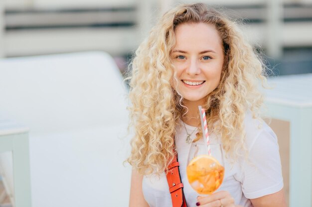 Positive curly young female with healthy clean skin dressed in casual white t shirt holds cold beverage looks happily at camera enjoys summer rest breathes fresh marine air outdoor at beach