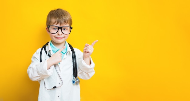 Medico carino positivo del ragazzo del bambino che indica con lo spazio vuoto dell'indice che mostra i migliori prezzi bassi. indossa l'uniforme medica isolata sullo sfondo di colore giallo