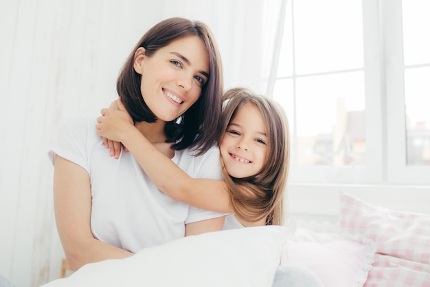 Positive daughter and mum embrace each other being in high spirit after good sleep pose in bedroom smile gently on face have pleasant appearance Pretty child with her mother Morning and bed time