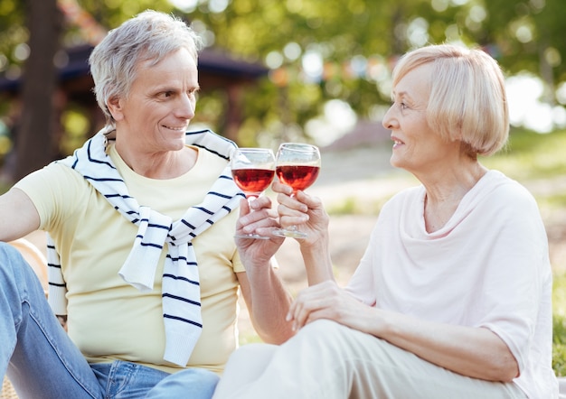 Positive delighted aging couple expressing delight while enjoying picnic and drinking wine outdoors