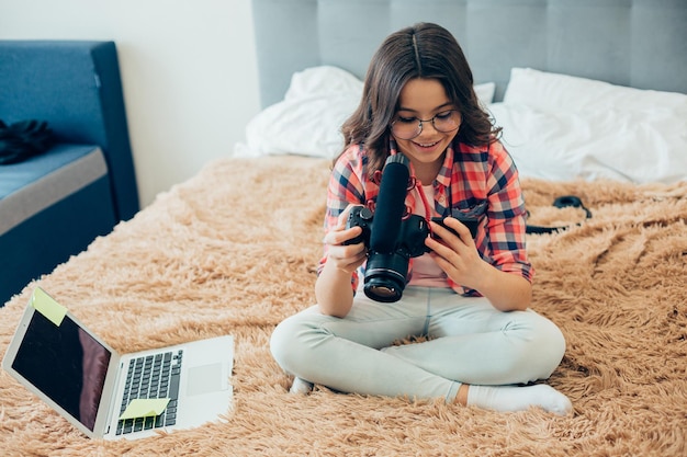 Positiva ragazza felice seduta sul letto con un laptop al suo fianco e guardando lo schermo di una telecamera