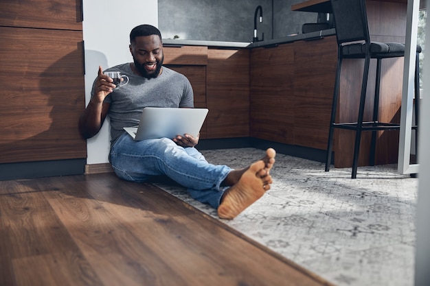 Positive delighted man holding cup with coffee in right hand while looking at screen of his laptop