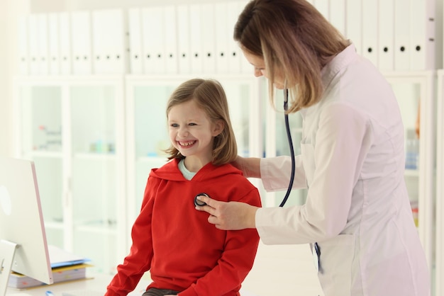 Positive doctor checks heart of cute little girl with stethoscope happy child smiles doing