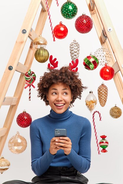 Positive dreamy woman with dark curly hair uses smartphone for\
sending messages of congratulations on new year wears reindeer\
horns and blue turtleneck poses around decorative toys on\
ladder