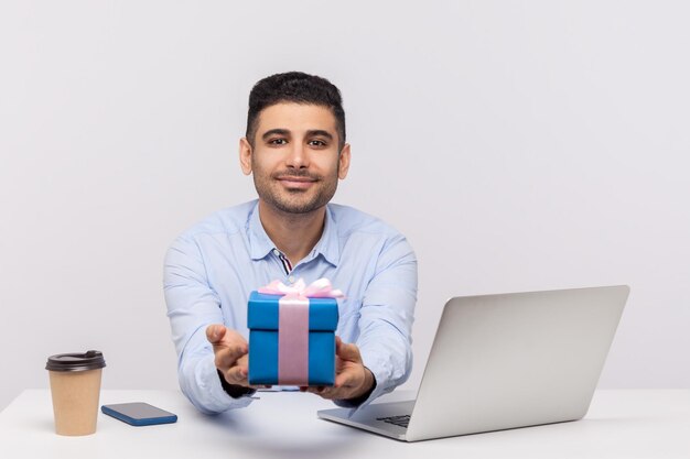 Positive elegant businessman sitting in office workplace giving gift box to camera and smiling friendly holding present bonus congratulating on professional holiday indoor studio shot isolated