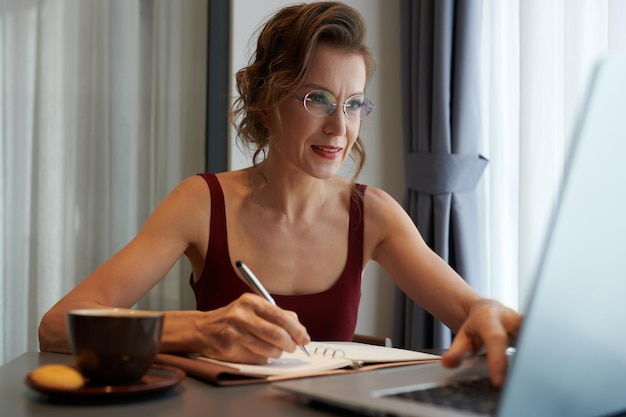 Фото Позитивная элегантная зрелая женщина в очках смотрит онлайн-курс на ноутбуке