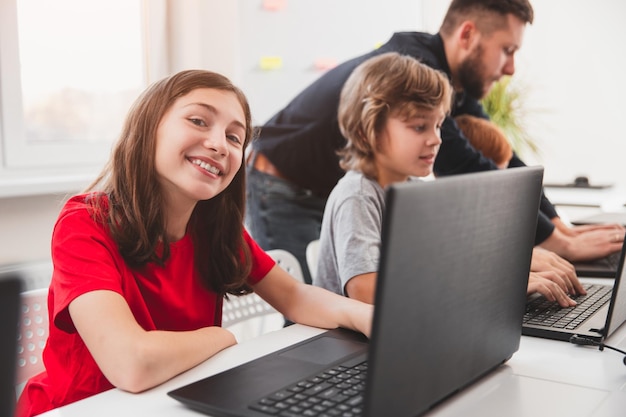긍정적인 초등학교 여학생은 반 친구들과 교사와 함께 교실에서 기술 수업을 하는 동안 노트북과 함께 책상에 앉아 웃고 카메라를 바라보고 있습니다.