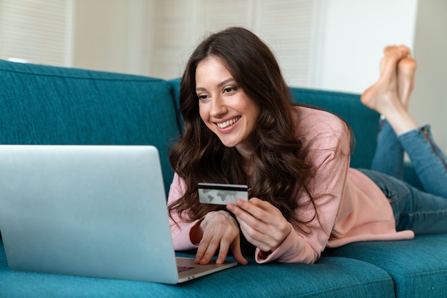 a positive emotional young woman indoors at home using laptop computer holding credit card on sofa.