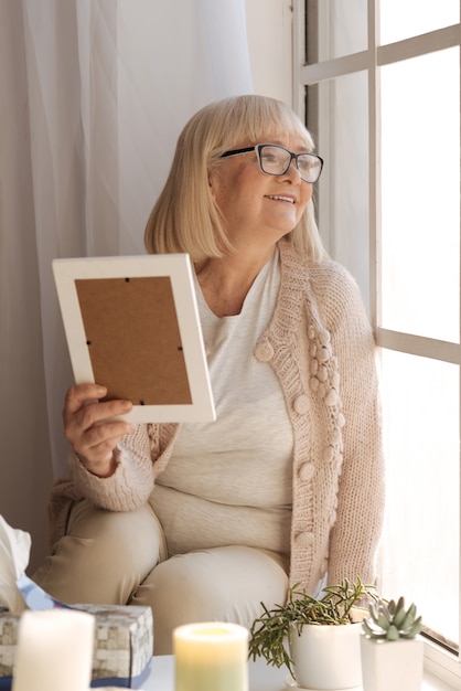 Positive emotions. happy cheerful elderly woman holding a photograph and looking into the window while being in a great mood