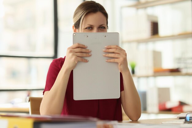 Positive employee hides face behind clipboard in company office closeup woman happy of