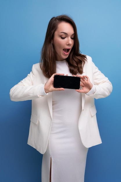 Positive energetic young brunette with long hair woman in white dress holding smartphone with mockup