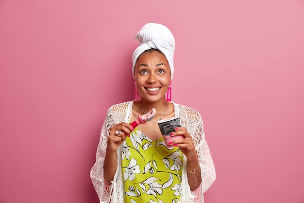 Positive ethnic housewife wears casual home clothes wrapped towel on head eats tasty cold ice cream with spoon looks above smiles toothily enjoys cold summer dessert isolated over pink wall