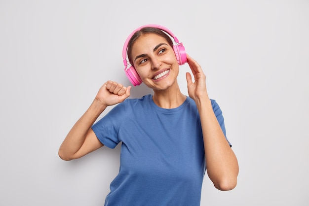 Positive european woman wears pink stereo headphones on ears
enjoys favorite song has cheerful dreamy expression dressed in
casual blue t shirt isolated over white background enjoys
playlist