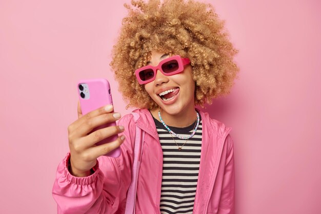 Positive fashionable woman with curly blonde hair sticks out\
tongue clicks selfie on smartphone makes photo of herself wears\
sunglasses and windbreaker isolated over pink background people and\
style