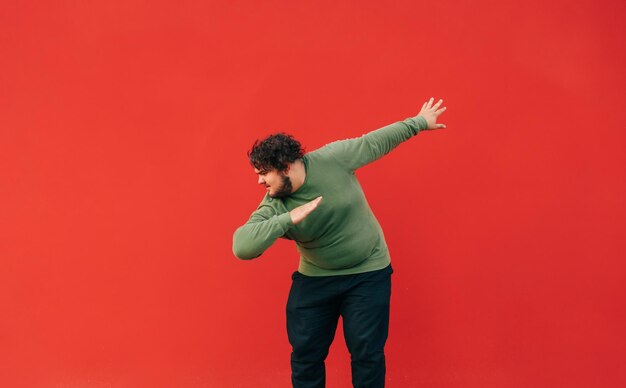 Positive fat man in casual clothes and curly hair dances a dub dance on a red wall background