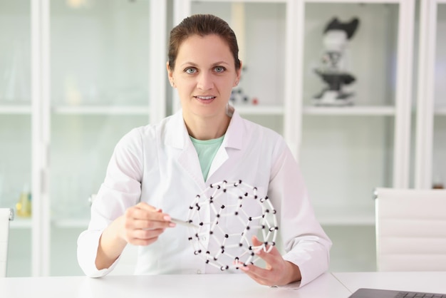 긍정적인 여성 화학자는 흰색의 여성을 손에 들고 있는 현실적인 분자 모델에 펜을 가리킵니다.