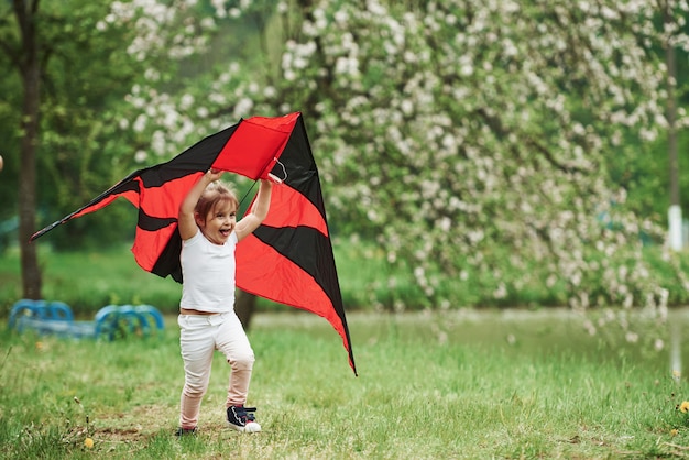 Positive female child running with red and black colored kite in hands outdoors