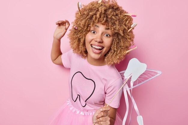 Positive female dental fairy dances carefree with magic wand
smiles broadly dressed in t shirt and skirt comes on costume party
entertains children smiles broadly isolated over pink
background